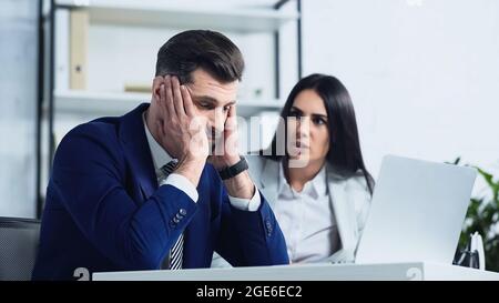Trauriger Geschäftsmann berührt den Kopf in der Nähe verschwommener Geschäftsfrau, die im Büro streitig ist Stockfoto