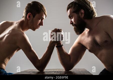 Stark muskulös bärtiger Mann, der einen schwach schwachen Mann am Arm kämpft. Arme ringen dünne Hand, großer starker Arm im Studio. Zwei Männerhände umklammten Arm-Wrestling Stockfoto