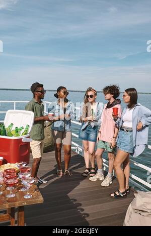 Junge Männer und Frauen feiern eine kleine Party auf dem hölzernen Pier, sie trinken Alkohol und plaudern Stockfoto