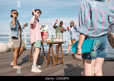 Eine Gruppe junger Leute freut sich, ihren Freund mit Skateboard auf der Party am Pier am See zu sehen Stockfoto