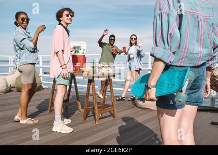 Junge Frau mit Skateboard kommt zur Party am Pier am See, wo Freunde Cocktails und Bier trinken Stockfoto