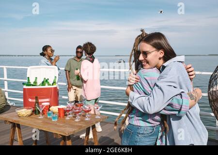 Fröhliche lächelnde junge Frau in Sonnenbrillen umarmte ihre Freunde, die sie auf der Geburtstagsfeier am Pier traf Stockfoto