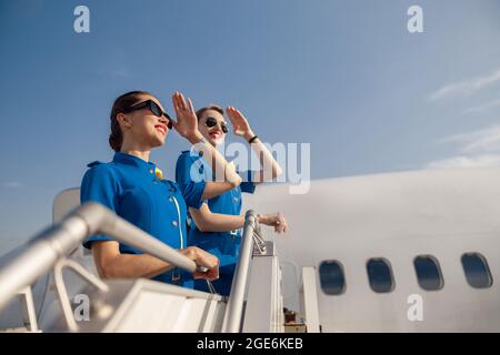 Portrait von zwei eleganten Luftverwaltungen in blauer Uniform und Sonnenbrillen, die die Augen mit der Hand bedecken und weit weg schauen, zusammen auf der Airstair stehen Stockfoto