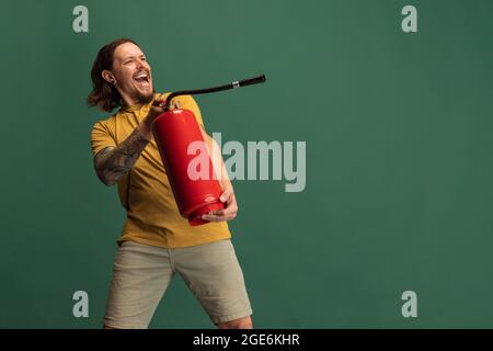 Comic-Porträt des jungen Mannes mit Feuerlöscher Spaß isoliert auf grünen Studio-Hintergrund. Konzept von Beruf, Arbeit, lustige Meme Emotionen Stockfoto