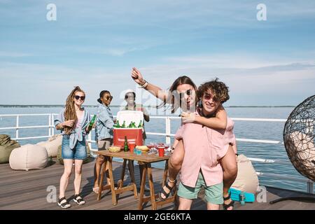 Hübscher junger Mann in Sonnenbrille, der seiner Freundin auf der Party auf dem hölzernen Pier eine Huckepack-Fahrt gab Stockfoto