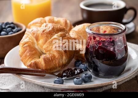 Croissants mit Heidelbeermarmelade und Heidelbeeren auf einem Teller mit Saft und Kaffee im Hintergrund Stockfoto