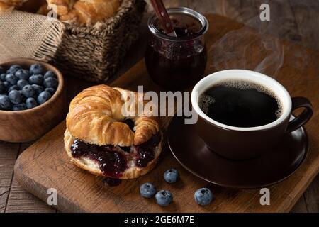 Croissant mit Heidelbeermarmelade und heißer Tasse Kaffee auf einem Holztisch Stockfoto