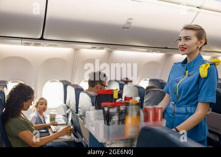 Weibliche Flugbegleiterin, die Passagieren im Flugzeug Essen serviert. Die Gastgeberin läuft mit dem Trolley auf dem Gang Stockfoto