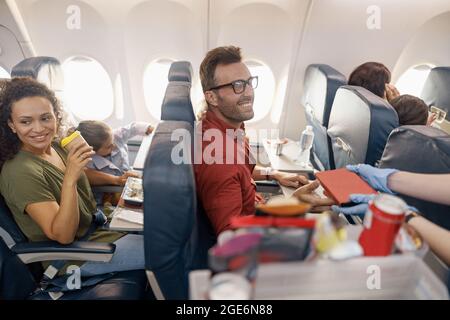 Glückliche Passagiere lächeln, während eine Flugbegleiterin das Mittagessen an Bord serviert Stockfoto
