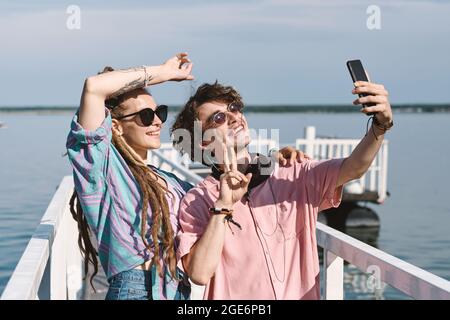 Glückliche junge Frau mit Dreadlocks und ihrem Freund im Lachshemd, die auf dem Pier steht und Selfie für Social Media macht Stockfoto