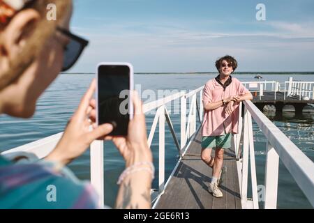 Junge Frau fotografiert ihren hübschen, stilvollen Freund in einer Sonnenbrille, die auf dem Pier posiert Stockfoto