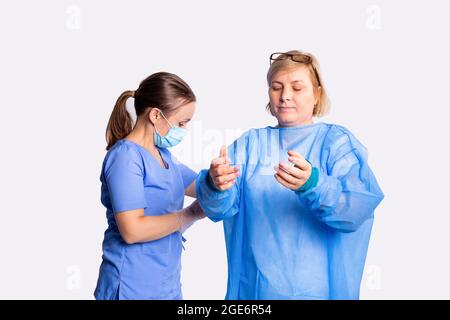 Die Krankenschwester in Schutzmaske, die einer blonden Frau mit einer Brille auf dem Kopf einen Schutzmantel für die Operation aufsetzt. Stockfoto