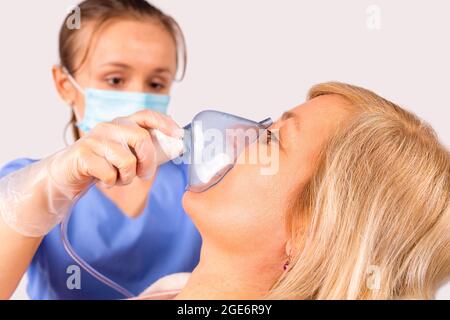 Die Ärztin in medizinischer Uniform und Schutzmaske, die der blonden Frau des Patienten eine Sauerstoffmaske zur Operation aufsetzt. Stockfoto