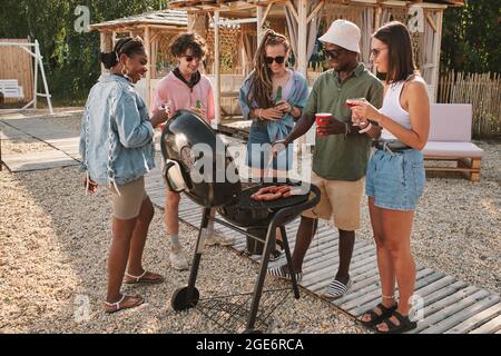 Diverse Gruppe von Freunden, die auf der Strandparty Hot Dogs auf dem Grill braten, trinken und reden Stockfoto