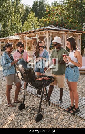 Freunde genießen Strandparty, sie machen gegrillte Hot Dogs, plaudern und trinken Bier Stockfoto