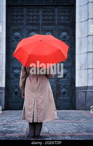 Eine Frau unter einem roten Schirm steht allein vor der Tür eines alten Hauses Stockfoto
