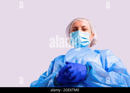 Foto von unten einer älteren Ärztin in einem medizinischen Kleid, einer Gesichtsmaske und blauen Schutzhandschuhen, die mit lächelnden Augen in die Kamera blicken. Stockfoto