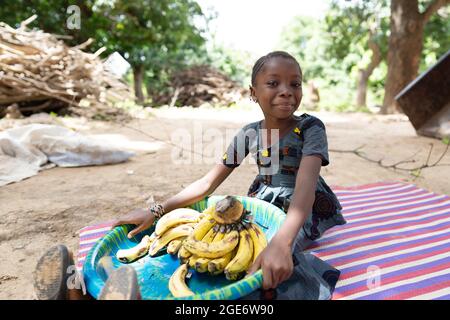 Hübsches kleines afrikanisches Mädchen, das einen großen Teller voller reifer Bananen auf den Knien hält und vor der Kamera grinst Stockfoto