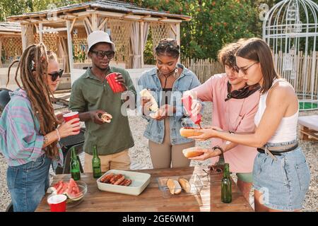 Diverse Freunde trinken Bier und essen Hot Dogs am Tisch am Strand und genießen eine kleine Sommerparty Stockfoto