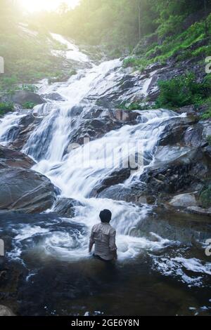 Ein Mann, der in fließendem Wasser steht, während er bei Sonnenaufgang den reinen Wasserfall in einem tiefen tropischen Regenwald betrachtet. Mae Wong Nationalpark, Thailand. Stockfoto