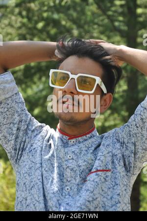 Porträt eines indischen hübschen jungen Mannes, der eine Sonnenbrille trägt und im Freien posiert, mit Händen auf dem Haar und seitlichem Blick Stockfoto