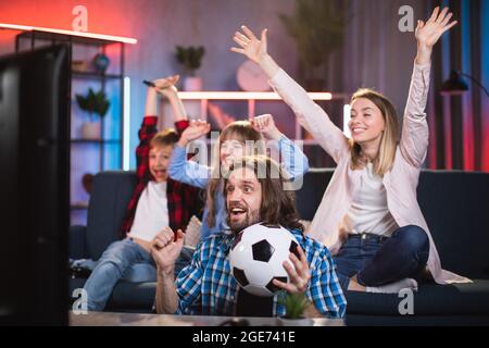 Emotionale kaukasische vierköpfige Familie sitzt auf der Couch und schaut sich Fußballspiele im Fernsehen an. Aufgeregt Eltern mit zwei Kindern unterstützen Lieblings-Team von zu Hause aus. Stockfoto