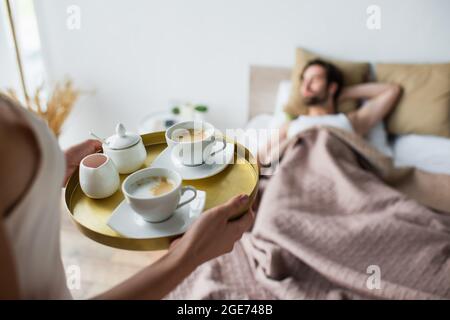 Frau hält Tablett mit Tassen Kaffee in der Nähe verschwommen und schlafenden Mann Stockfoto