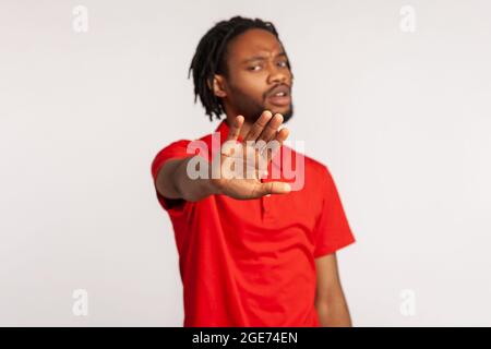 Mann mit Dreadlocks trägt ein rotes T-Shirt im lässigen Stil, mit einem ernsten Gesicht, das mit seiner Handfläche eine Stoppgeste zeigt und gegen Mobbing und Gewalt ist. Indoo Stockfoto