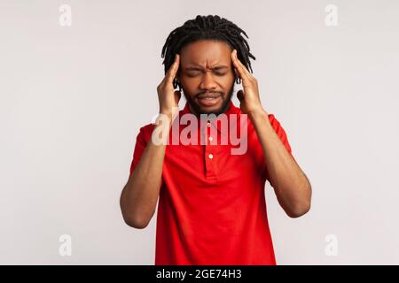 Attraktiver bärtiger Mann mit Dreadlocks, der ein rotes T-Shirt im lässigen Stil trägt und unter schrecklichen Kopfschmerzen leidet, das Gesicht runzelnd und die Finger auf den Schläfen hält. I Stockfoto