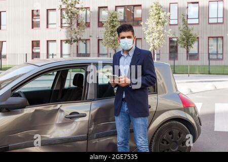 Junge Erwachsene Brünette Mann in chirurgische medizinische Maske, Jacke und Jeans posiert auf der Straße der Stadt mit digitalen Tablet in den Händen in der Nähe zerschmetterte Auto mit de Stockfoto