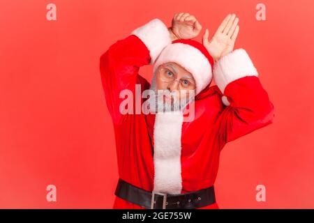 Lustiger älterer Mann mit grauem Bart, der das weihnachtsmannkostüm trägt, pout Lippen spielerisch und zeigt Hasenohren, die mit komischer kindlicher Grimasse schnaufeln. Innen Stockfoto