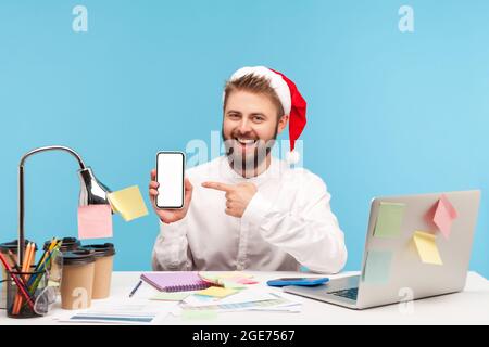 Lächelnder, positiver Mann im Büro des weihnachtsmannes zeigte auf das Smartphone mit leerem Display, das mit einem Laptop am Arbeitsplatz sitzt und Geschenke kauft Stockfoto