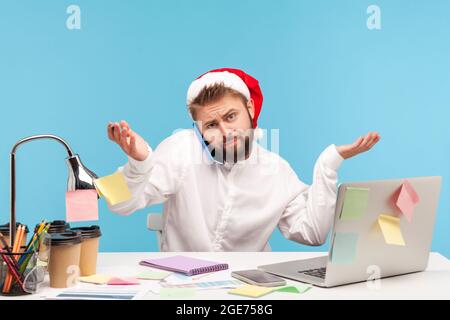 Verwirrt Mann in weihnachtsmann hat zuckende Schultern und die Hände heben Telefon reden, sitzen am Arbeitsplatz mit Laptop, arbeiten an Feiertagen, Frist. Stockfoto