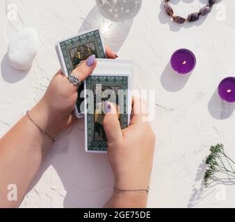 Hände mit violetten Nägeln und Ringen halten Tarot-Karten auf weißer Oberfläche mit Kristallkugel, Kerzen und Steinen. Draufsicht Stockfoto