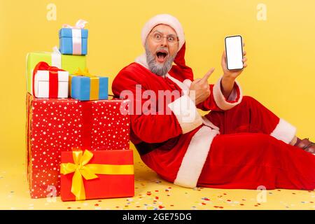Staunender älterer Mann mit grauem Bart, der ein weihnachtsmann-Kostüm trägt, sitzt auf einem weißen, leeren Bildschirm mit einem Smartphone, umgeben von Weihnachtsgeschenken Stockfoto