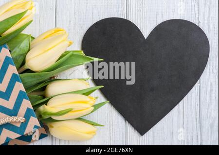 Frühlingskarte mit gelben Tulpen in Kraftpackung und herzförmiger Kreidetafel auf Holztisch. Platz für Text. Landhaus rustikalen Stil. M Stockfoto