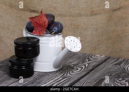 Große Pflaumen in einer Gießkannen aus Metall. Auf Kiefernbrettern. In der Nähe befindet sich ein Glas Marmelade. Pflaumen ernten. Stockfoto