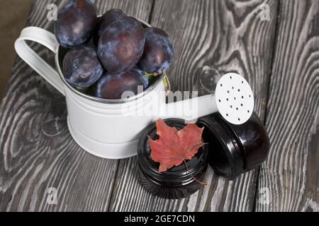 Große Pflaumen in einer Gießkannen aus Metall. Auf Kiefernbrettern. In der Nähe befindet sich ein Glas Marmelade. Pflaumen ernten. Stockfoto