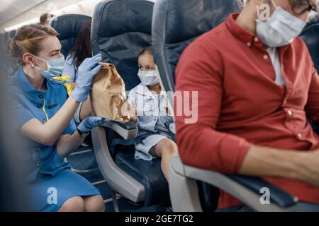 Mädchen in schützender Gesichtsmaske, die eine Flugbegleiterin ansieht, die dem kleinen Passagier das Mittagessen serviert Stockfoto