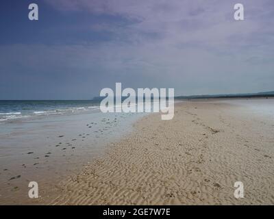 Ein riesiger, leerer, welliger Sandstrand mit Fußabdrücken, die in die Ferne und zum Hunt Cliff am Horizont führen, alles unter einem trüben, blauen Himmel. Stockfoto