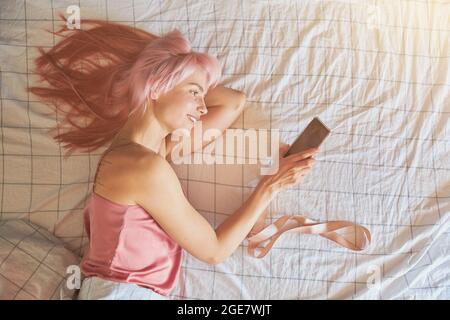 Lächelnde Frau schaut auf das Mobiltelefon, das auf dem bequemen Bett in der oberen Ansicht liegt Stockfoto