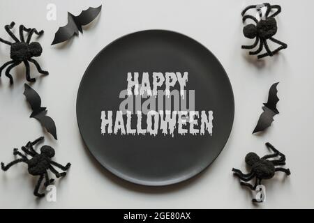 Halloween Einladung zur lustigen Horror Party. Schwarze Spinnen, Fledermaus und schwarzer Teller mit Text 'Happy Halloween' auf grauem Tisch. Flach liegend, Draufsicht. Stockfoto