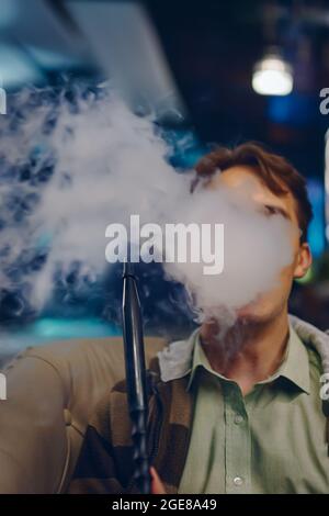 Mann im Café raucht Shisha. Starker Rauch von Shisha. Junger Mann auf dem Sofa im Restaurant. Farbige Lichter auf dem Hintergrund. Entspannen Sie sich in der Shisha Bar. Stockfoto