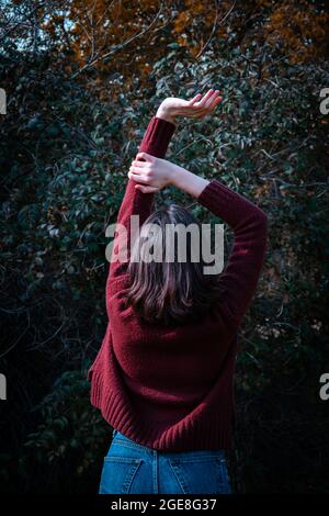 Happy junge Brünette Mädchen in einem braunen Pullover stehen mit Heben ihre Hände in einer Waldlandschaft. Rückansicht, vertikaler Rahmen, Tonung. Stockfoto