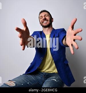 Porträt eines lachenden erwachsenen Mannes in zerrissenen Jeans, blauer Jacke und gelbem T-Shirt, der seine Hände aushält und uns umarmen wird Stockfoto