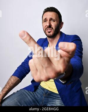 Porträt eines unglücklichen, irrigen, unrasierten erwachsenen Mannes in einer lässigen blauen Jacke und einem gelben T-Shirt, das den Mittelfinger aushält Stockfoto