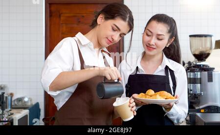 Der junge kaukasische Barista gießt Milch aus dem Krug in eine Papierkaffeebecher. Asiatische Assistentin in einer Schürze, die einen Croissantteller hält, steht neben ihr. Morn Stockfoto