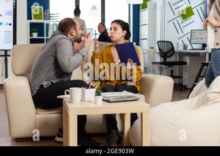 Geschäftskollegen streiten sich, während der Arbeitszeit auf der Couch sitzend, sich gegenseitig in die Wange nehmen, während verschiedene Kollegen im Hintergrund Angst vor der Arbeit haben. Mitarbeiter, die aufgrund von Fehlern aus dem Projekt Konflikte haben Stockfoto
