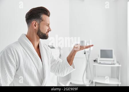 Stilvoller bärtiger Mann im Bademantel präsentiert Cremedose im Schönheitssalon Stockfoto