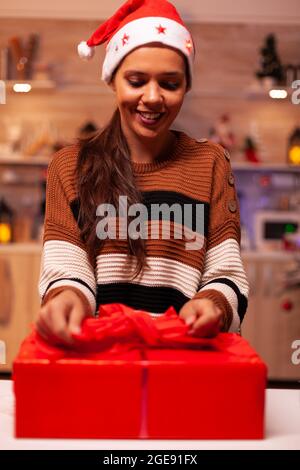 Traditioneller Erwachsener mit weihnachtsmütze mit Band zum Binden der Schleife auf der weihnachts-Geschenkbox für Freunde im festlichen Zuhause. Junge Frau Dekoration Geschenk mit Geschenkpapier für Feiertagsfeier Stockfoto